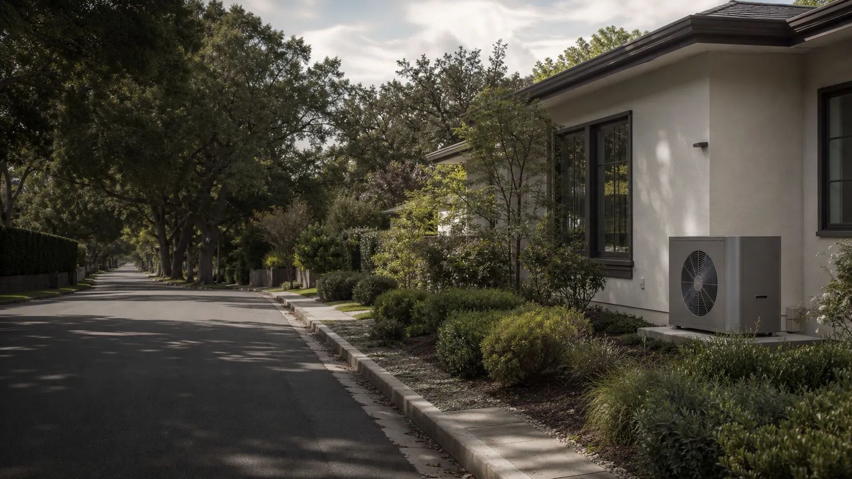 Tree-lined Burlingame residential street with Spanish-style stucco homes and a modern outdoor heat-pump unit beside one house, coastal hills in the distance.